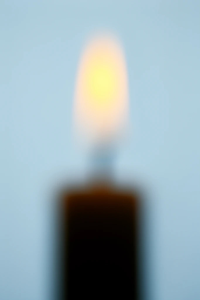A heavily out-of-focus lit candle against a smooth, pale blue background. A dark cylindrical silhouette anchors the frame beneath a soft, luminous yellow-orange flame. Extreme bokeh creates a minimalist composition of smooth gradients and color contrast.