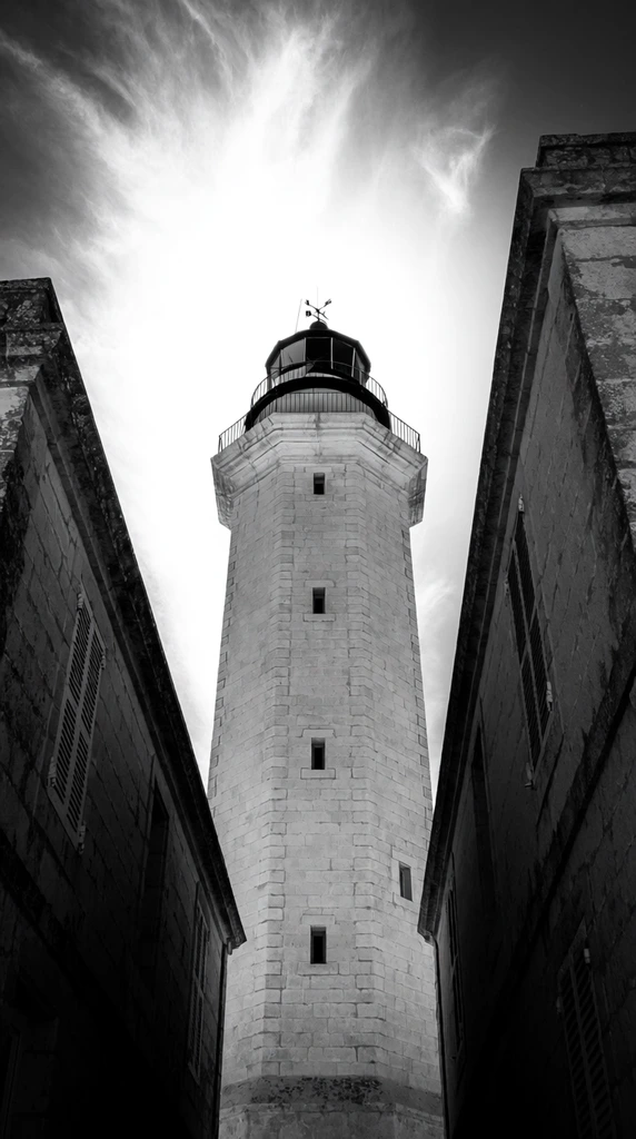 A towering octagonal stone lighthouse photographed from a dramatic low angle in stark high-contrast black and white, framed tightly by adjacent historic buildings. The bright central tower features a dark lantern room encircled by a metal railing deck, crowned with a weather vane, and punctuated by a vertical column of small recessed rectangular windows. It stands wedged between heavily shadowed, weathered stone structures with shuttered windows flanking both sides. The severe upward perspective emphasizes the imposing architectural scale of the lighthouse against an intensely backlit sky swept with bright, wispy cirrus clouds.