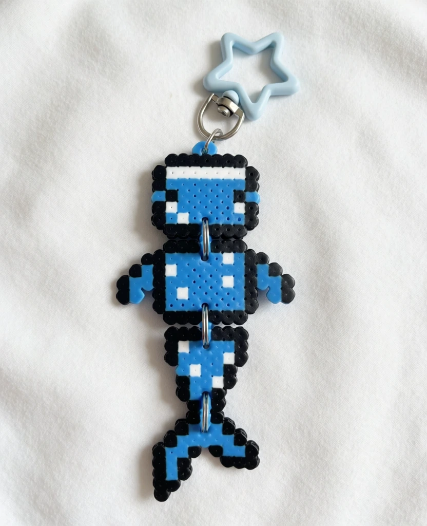 A blue whale shark keychain made of fused plastic beads is positioned vertically in the center of the frame against a white fabric background. The pixelated figure is divided into three distinct segments—head, body, and tail—which are joined together by small silver metal rings, allowing for articulation. The whale shark is primarily bright blue with small white square spots and a thick black outline. The head segment features a white bar at the top and two small white squares for eyes. The torso segment has several white spots and two small blue fins protruding from the sides. The tail segment tapers down to a forked fin and also contains white spots. At the top of the head, a silver metal jump ring connects to a lobster claw clasp, which is also attached to a pale blue, star-shaped plastic charm. The entire object lies flat on a soft, white, slightly wrinkled textile surface. The lighting is bright and even, casting subtle shadows to the left, and the camera angle is a direct overhead shot, capturing the detail of the ironed beads and the texture of the fabric.
