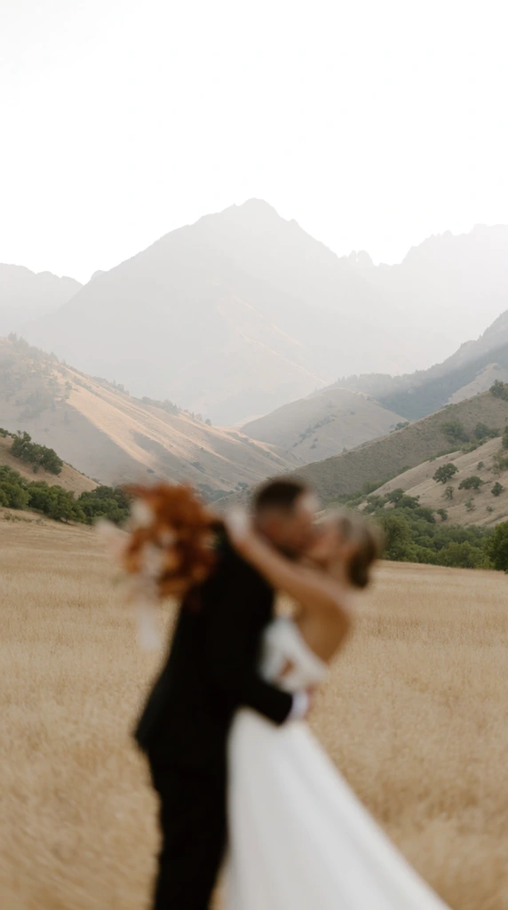 An intentionally blurred bride and groom kissing in a dry golden meadow, contrasting against a vast, sharper hazy mountain range. The groom in a black suit embraces the bride in a white dress holding a rust-colored floral bouquet. Rolling foothills with green foliage rise into towering peaks fading into an overexposed white sky. This romantic photograph features a striking focal inversion, leaving the foreground couple obscured by soft bokeh to emphasize the majestic, atmospheric landscape behind them in a muted color palette of tan, sage, and pale gray.