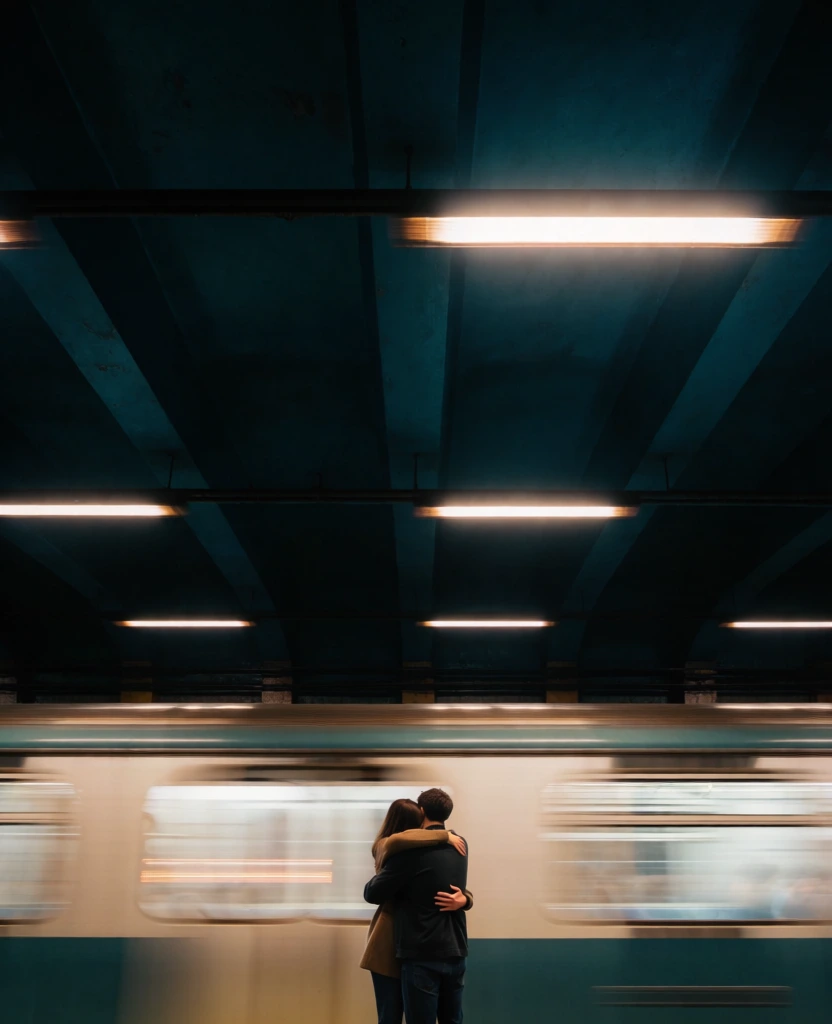 A tender, cinematic moment on a subway platform shows two people embracing tightly while a train rushes past in a blur, making the stillness of their hug feel intimate against the speed of the city. photographic image, long-exposure street photography, cinematic realism, urban romance, fine-art portraiture, moody, melancholic, intimate, nocturnal, atmospheric, soft-focus motion blur, medium-high visual complexity, Wong Kar-wai adjacent, Christopher Doyle adjacent, Gregory Crewdson adjacent, teal-and-amber palette, muted olive, pale cream, smoky blue, deep black, low-key exposure, luminous highlights, soft but dramatic contrast, shallow to moderate depth of field, likely slow shutter speed, possible handheld or stabilized shot, pronounced horizontal motion streaks from passing train, slight glow around lights, no obvious digital noise. The frame is vertical and dominated by darkness at the top, where the station ceiling or tunnel area forms a large black-green field. Across the upper third, faint horizontal lines suggest cables or structural elements, and two blurred rectangular station signs hover in the darkness: one left of center glowing orange with unreadable text, another on the upper right glowing pale greenish-white. Cutting across the middle of the image, the subway train becomes bands of cream, silver, and blue-green smeared horizontally from left to right, its windows and lights stretched into luminous stripes. In the lower center-right foreground stand the couple, seen mostly from behind. The taller person, likely with dark hair tied in a rounded bun, wears a long dark overcoat with a broad back and soft folds; their right arm wraps around the other person’s back. The shorter person leans their head into the taller person’s chest and shoulder, their long auburn-brown hair catching the light. They wear a light, flowing dress or coat in off-white or pale gray, the fabric draping downward in soft vertical folds. In their left arm, near the lower left of the embrace, they hold a bouquet wrapped in translucent paper; the flowers appear cream or white with darker dried accents and greenery. The shorter person’s right hand rests around the taller person’s waist at the right edge of the embrace. The couple is still, centered slightly right, while the train streaks behind them, turning the scene into a visual contrast between motion and devotion.