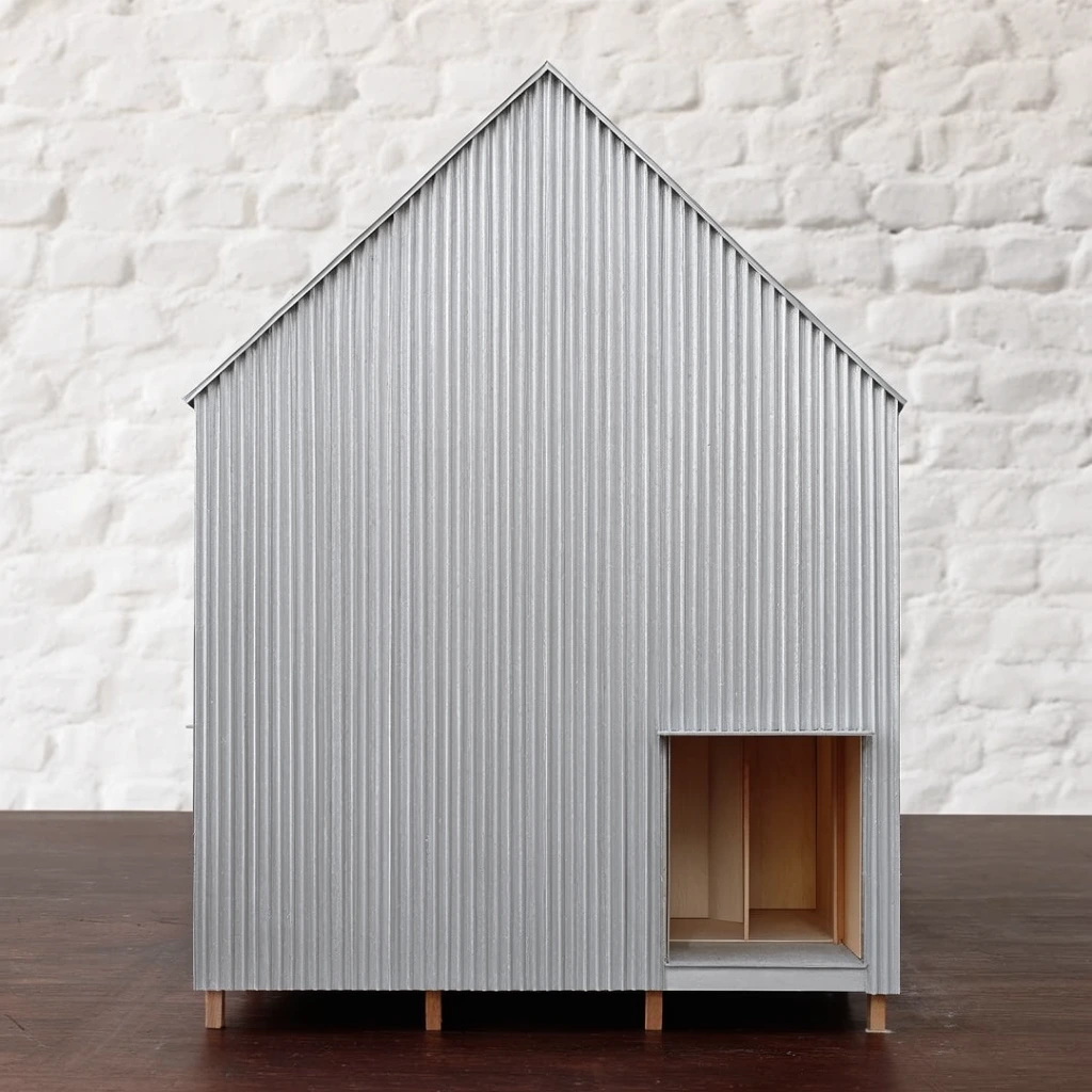 architectural scale model of a minimalist house, dramatically tall gable roof, exterior entirely clad in light silver-gray vertical corrugated metal, elevated on short wooden stilts, rectangular recessed entryway on the lower right revealing wooden interior panels, flat front elevation perspective, resting on a dark wood surface, textured white painted brick wall background, soft diffused lighting, precise vertical lines, clean geometry