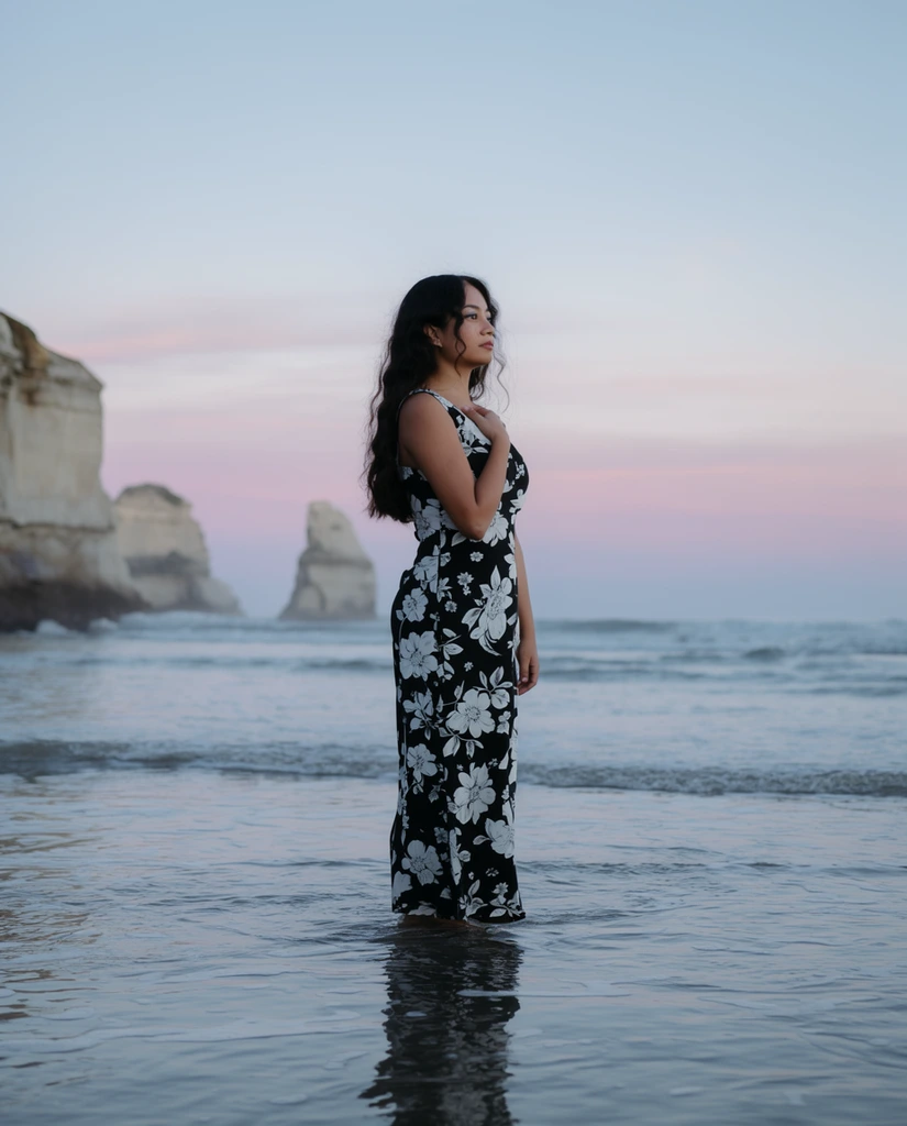 A young woman stands ankle-deep in gentle ocean waves against a serene twilight horizon. She has long, dark, wavy hair and wears a sleeveless, floor-length black dress adorned with large, contrasting white floral patterns. Her right hand rests delicately on her upper chest, and her face is turned in profile, gazing calmly out of frame. The calm sea surrounds her lower legs, reflecting her silhouette in the shimmering shallow water. In the distant background, sheer-sided rocky sea stacks rise abruptly from the ocean. The sky above transitions smoothly from pale blue to soft pastel pinks and lavenders. Captured as a cinematic portrait photograph with a slightly shallow depth of field, the soft diffused dusk lighting emphasizes cool, muted coastal tones.