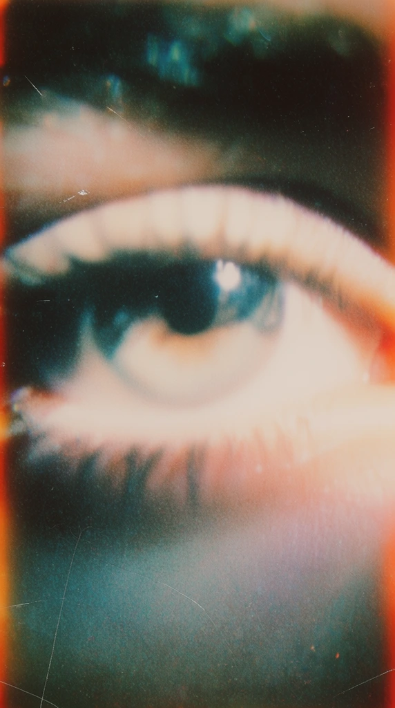 A heavily grained, out-of-focus vintage instant photograph of a close-up of a single eye. The eye dominates the composition, its pupil, iris, and eyelashes rendered with a severe soft focus and a dreamy, ethereal blur. Milky, warm peach and pale cream highlights wash over the center of the eye, contrasting starkly against deep, crushed cyan and dark teal shadows blending into the surrounding skin. The image is saturated with physical lo-fi film artifacts, including prominent dust specks, thin white hairline scratches scoring the surface, and intense, chunky film grain. Vibrant orange and red light leaks flare along the extreme edges of the frame. A heavy, murky vignette darkens the periphery, amplifying the nostalgic, degraded Polaroid aesthetic characterized by subtle chromatic aberration, low-resolution textures, and a faded, moody emulsion.