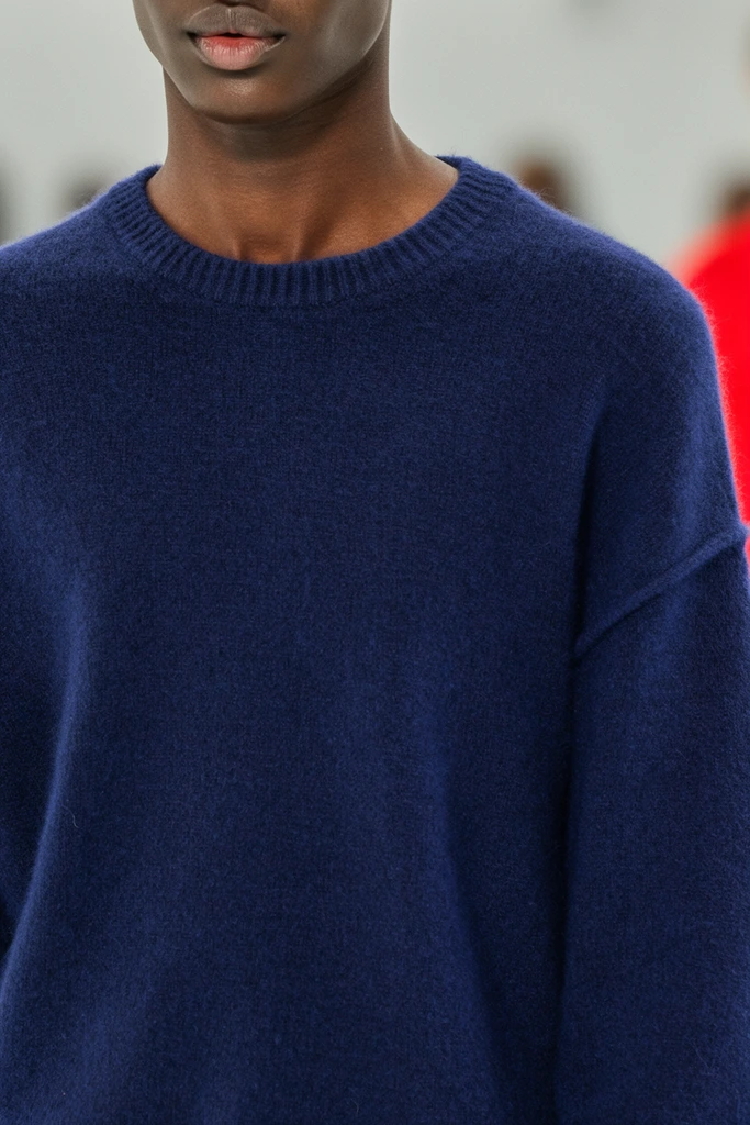 A tight close-up fashion portrait focusing on the chest, neck, and lower face of a dark-skinned model wearing a solid navy blue knit sweater. The garment features a soft woolen texture, a ribbed crew neckline, and a dropped shoulder seam. The model has relaxed, slightly parted lips and a defined jawline. The shallow depth of field sharply captures the textile fibers and skin texture against a blurred light grey background, which is accented by an out-of-focus patch of vibrant red on the right edge. Even, bright lighting highlights the fuzziness of the sweater and the smooth complexion, creating a clean, highly detailed editorial composition.
