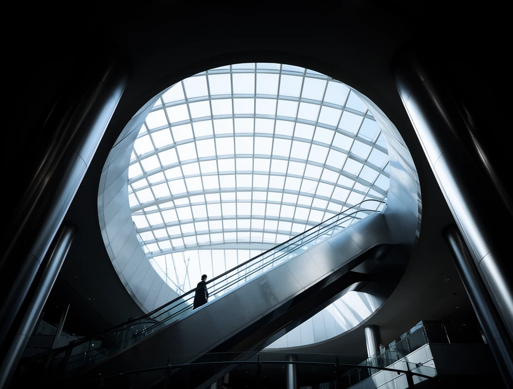 Modern architectural interior, dramatic low angle looking up, massive circular ceiling cutout framing a curved grid glass roof, bright cool daylight, ascending escalator crossing the central opening, solitary silhouetted figure riding upwards, thick metallic cylindrical columns, glass guardrails, high contrast lighting, deep foreground shadows, cool silver and black palette, geometric composition