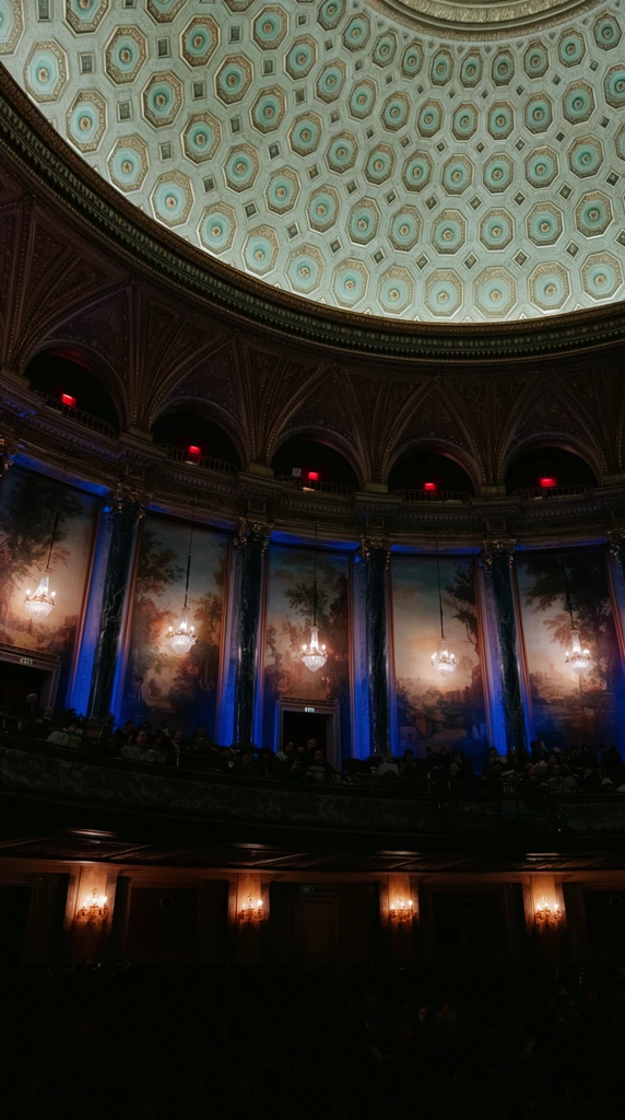 A grand atmospheric theater interior viewed from a low angle focusing on a magnificent domed ceiling adorned with pale teal and gold octagonal coffers. Intricate ribbed vaulting meets a side wall lined with dark marble columns framing classical painted landscape murals. Glowing crystal chandeliers hang before the murals. Deep blue accent lighting washes the pillars, contrasting with small red lights inside upper architectural arches. A curved balcony holds a silhouetted audience, while lower seating tiers fade into deep shadow pierced by warm amber sconces, defining a richly textured architectural space.