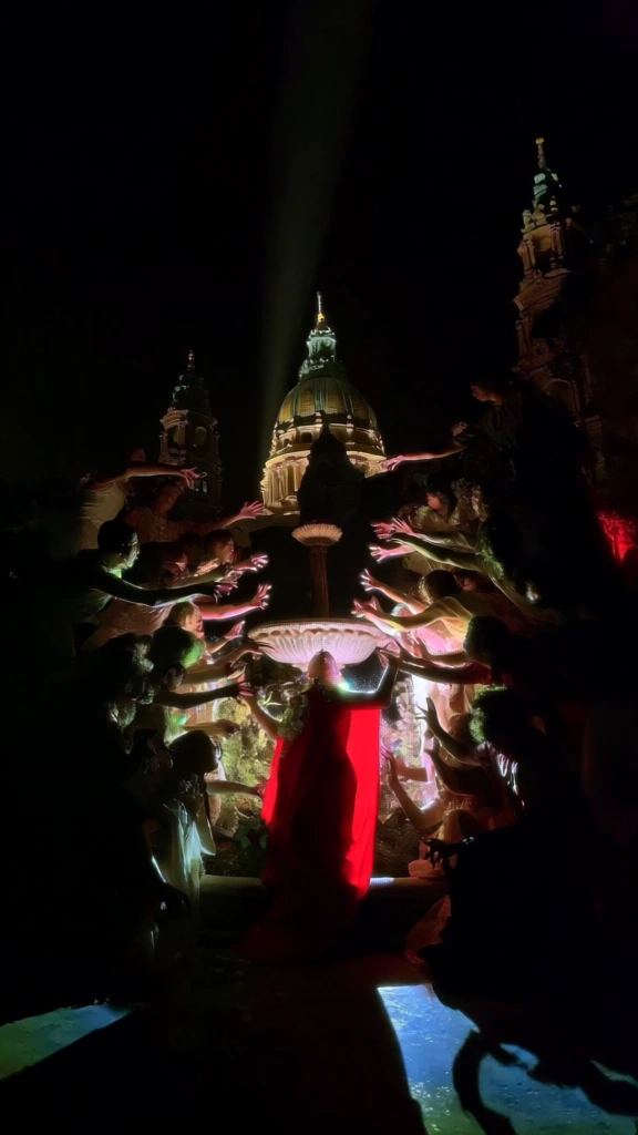 cinematic theatrical chiaroscuro photography, high-contrast scene at logan circle in philadelphia, centripetal composition with figures reaching mystical hands towards a central figure in a red cloak standing before the swann memorial fountain, the majestic cathedral basilica dome glowing in a singular beam of light, deep pitch-black shadows, moody ritualistic atmosphere, vibrant pops of red against dark gothic tones, low-angle perspective, immersive drama, grand scale, grain texture