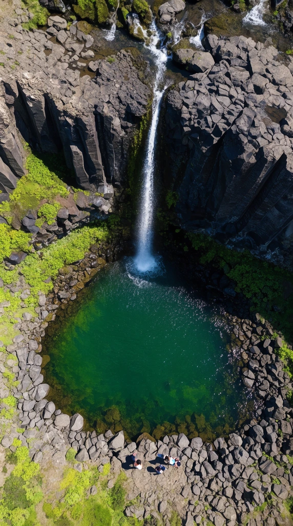 Aerial top-down view of a narrow waterfall cascading over dark basalt cliffs into a deep emerald-green plunge pool. Jagged grey rocks and patches of bright green vegetation surround the vibrant basin. Tiny human figures stand on the bottom rocky edge, providing scale to the sunlit geological formation.