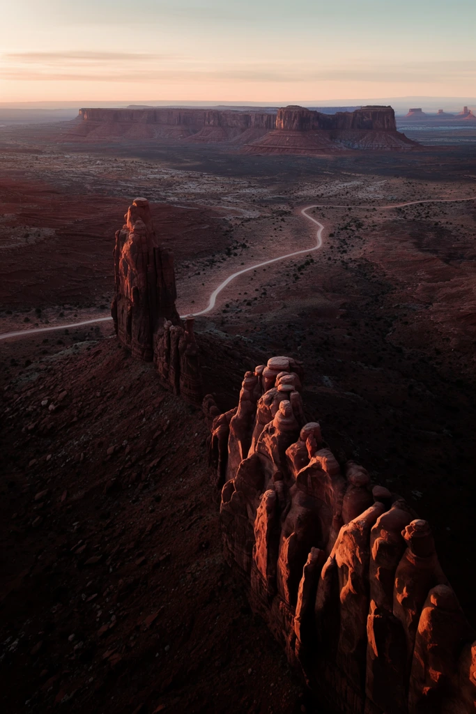 A vast desert landscape at dawn features towering red rock formations and a winding dirt road. A jagged sandstone ridge and a tall rock spire dominate the foreground, their sunlit edges glowing deep reddish-brown against the shadowed valley. Below, a pale trail snakes through the arid basin. Distant flat-topped mesas stretch toward the horizon under a soft peach and pale blue sky. This high-angle aerial photograph captures dramatic golden hour lighting, deep contrast, and stark geological textures.