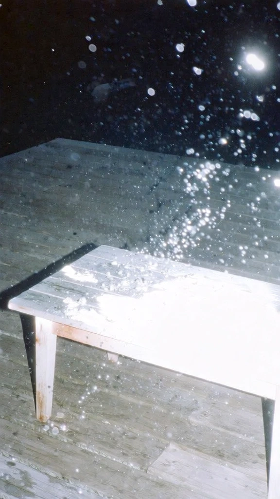 high-intensity direct flash photography, night scene on a weathered wooden deck featuring a minimalist coffee table, harsh overexposed white highlights hitting the wood, deep black grainy shadows in the background, shimmering light particles frozen in mid-air like crystalline droplets, cool blue and silver color palette, raw snapshot aesthetic, cinematic film grain, minimalist off-center composition, lo-fi fashion editorial style, high contrast, ethereal luminescence.