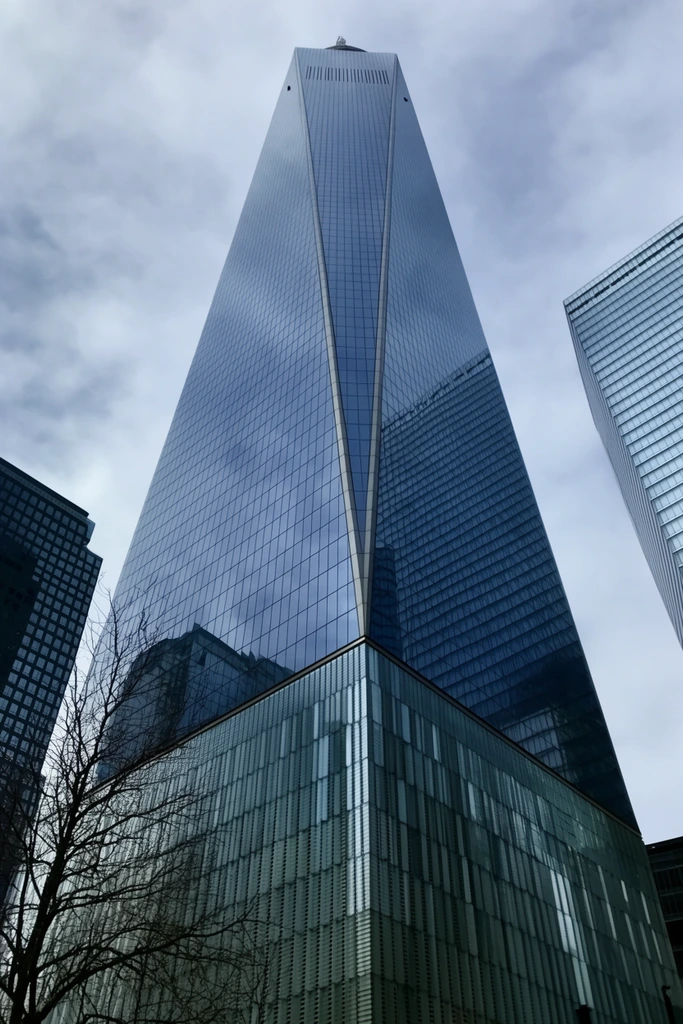 A towering modern glass skyscraper captured from a sharp low angle dominates an overcast urban sky. The crystalline structure features massive triangular facets tapering toward the peak, its steel-blue reflective surface mirroring the clouds and adjacent grid-patterned high-rises. Textured vertical metallic slats detail the building's base. Bare tree branches stretch into the lower left foreground, contrasting with the sleek architecture, while darker, box-like buildings frame the central monument to emphasize its colossal scale and converging lines.