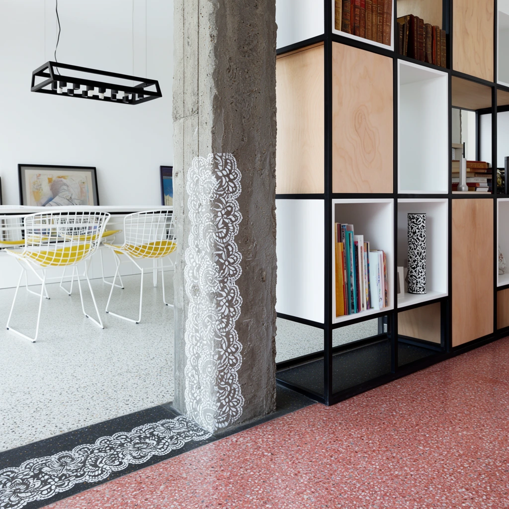 bespoke shelving unit acting as a room divider, central rough grey concrete pillar decorated with delicate white lace stencil patterns, black metal framing alternating with light plywood panels and white open cubbies, bound volumes and a patterned cylinder on shelves, foreground reddish-pink speckled terrazzo flooring, midground light grey speckled terrazzo, dark transition strip with matching white lace stencils, background bright white dining area, black rectangular chandelier, white table, white wire chairs with yellow accents, framed artworks leaning against a white wall, clean architectural photography, natural daylight, sharp focus, geometric composition