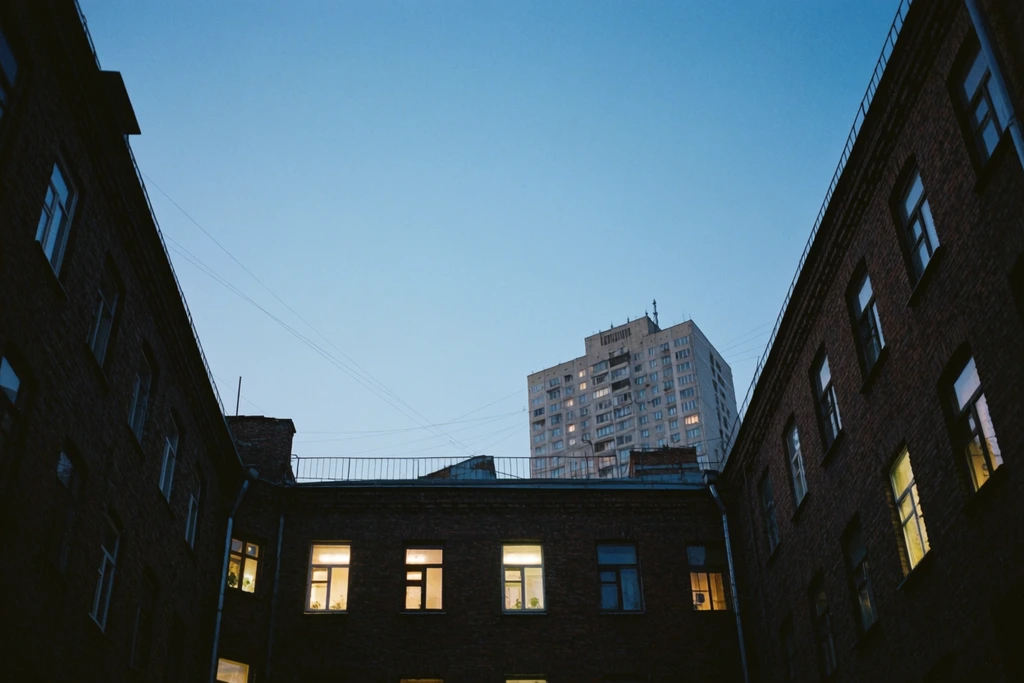 low angle view of an urban courtyard at dusk, dark brick buildings framing a clear deep blue sky, warm yellow lit windows, distant high-rise, film grain
