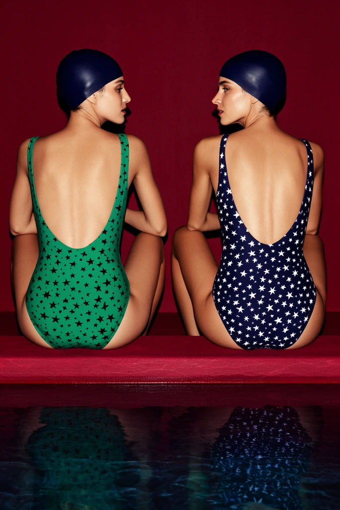 Two women in patterned swimsuits and dark swim caps sit symmetrically on a crimson ledge overlooking a dark pool. The left woman wears a vibrant green low-back swimsuit with black stars, turning her head right in profile. The right woman wears a dark blue low-back swimsuit with white stars, turning her head left in profile. Both wear smooth navy swim caps. They rest with knees bent outward against a solid, deep red background. Below, the dark water reflects the red ledge and their lower bodies. The composition relies on striking symmetry, captured with harsh, direct lighting that casts sharp shadows and highlights the rich, saturated palette of crimson, emerald, and navy in a polished editorial fashion aesthetic.