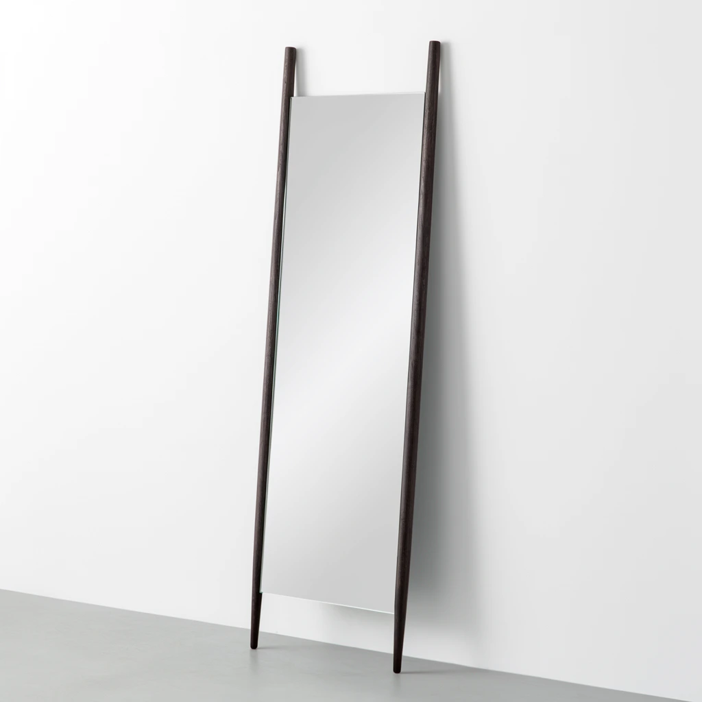A minimalist full-length leaning floor mirror rests against a stark white wall on a smooth light grey floor. A tall rectangular pane of reflective glass is suspended between two slender, dark espresso-colored wooden vertical poles. These textured uprights serve as the only framing, leaving the top and bottom horizontal edges of the glass exposed. The side poles extend above the top edge of the mirror and taper downward to form resting feet. Soft, diffused studio lighting creates a smooth white gradient reflection on the glass surface and casts a subtle drop shadow on the wall behind the right side. The three-quarter perspective highlights the angled posture and sharp geometric contrast of the modern design within the bright, pristine environment.