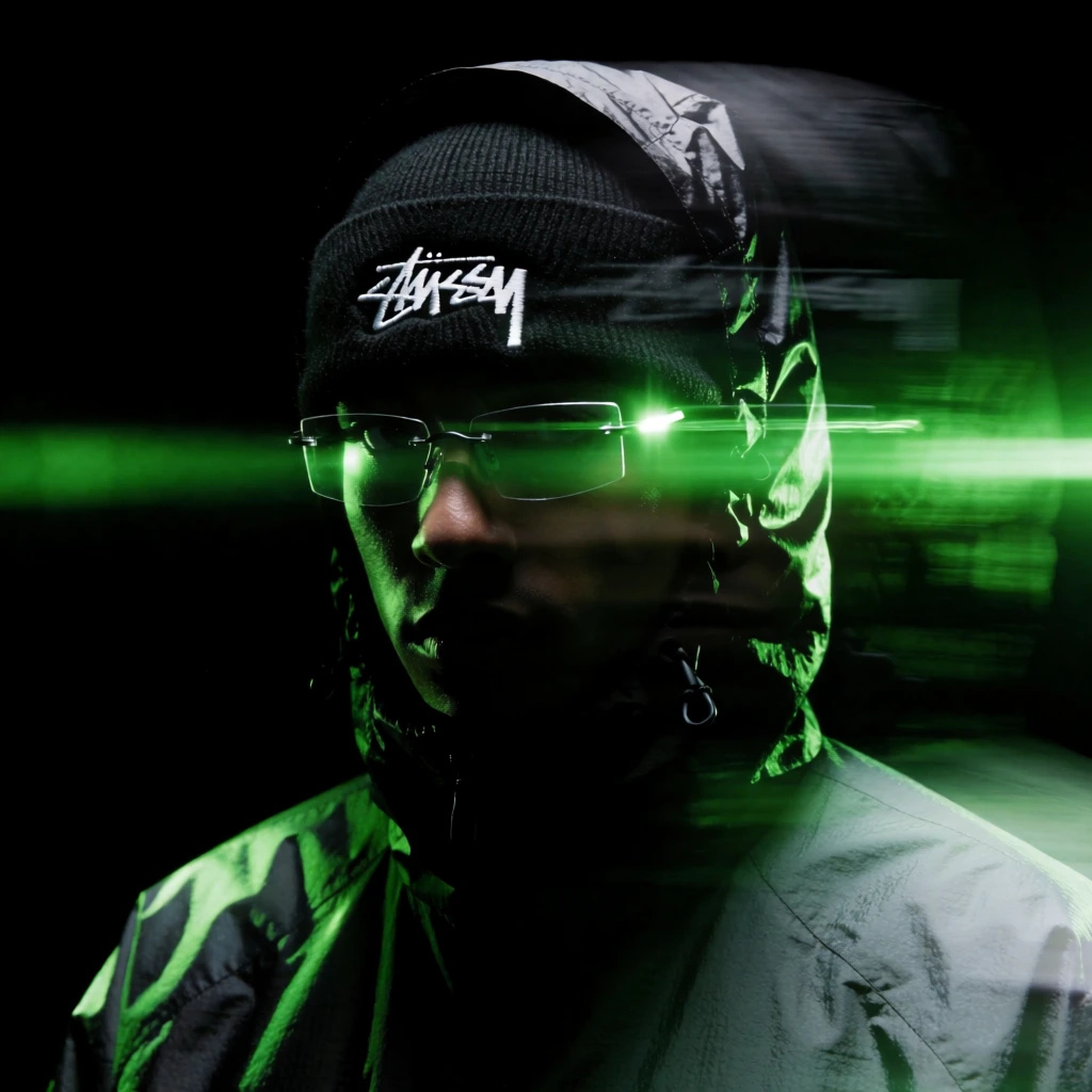 A person wearing a black beanie and a reflective hooded jacket is depicted in a high-contrast, green-tinted portrait featuring prominent motion blur and digital glitch effects. The central figure is shown from the chest up, with their head tilted slightly downward. They are wearing a black knit beanie that features the "Stüssy" logo in its signature white script font, centered on the forehead. Over the beanie, a dark, shiny hooded jacket is worn with the hood pulled up, its material reflecting the light in sharp, angular highlights. The person wears thin-rimmed rectangular glasses that catch vibrant emerald green light along their edges. The right side of the frame is heavily stylized with horizontal motion blur and light streaks that stretch the subject's features and the logo across the dark space. The background is a solid, deep black, providing no environmental context and focusing entirely on the subject. Lighting is low-key and directional, coming from the upper right, which casts deep shadows over the face while illuminating the left side and the bridge of the nose with a saturated green glow. The entire image has a gritty, grainy texture with visible digital noise, contributing to a techno-aesthetic or cinematic style. The color palette is strictly limited to dark blacks and various shades of luminous green.