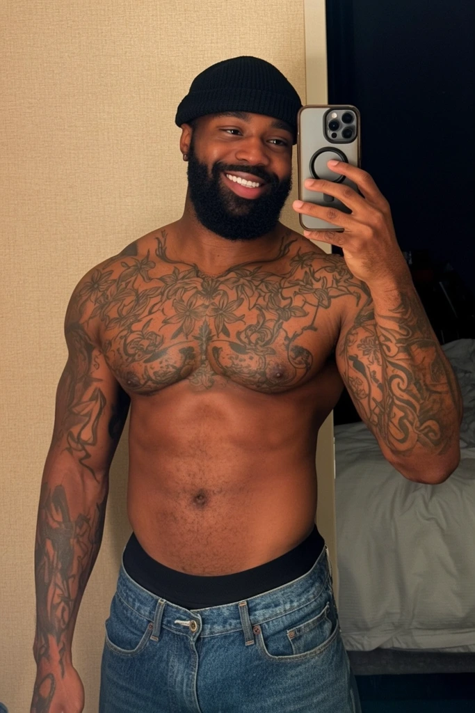 A muscular, shirtless Black man smiles brightly while taking a mirror selfie. He features a full beard, a black knit beanie, and low-slung blue denim jeans revealing a thick black underwear waistband. Intricate black-ink floral and abstract tattoos densely cover his chest and arms. He holds a smartphone with a dark circular ring grip. The reflection captures a textured beige wall alongside a dark doorway opening into a room with a white-linened bed. Warm indoor lighting highlights the contours of his torso and striking body art in this candid waist-up portrait.