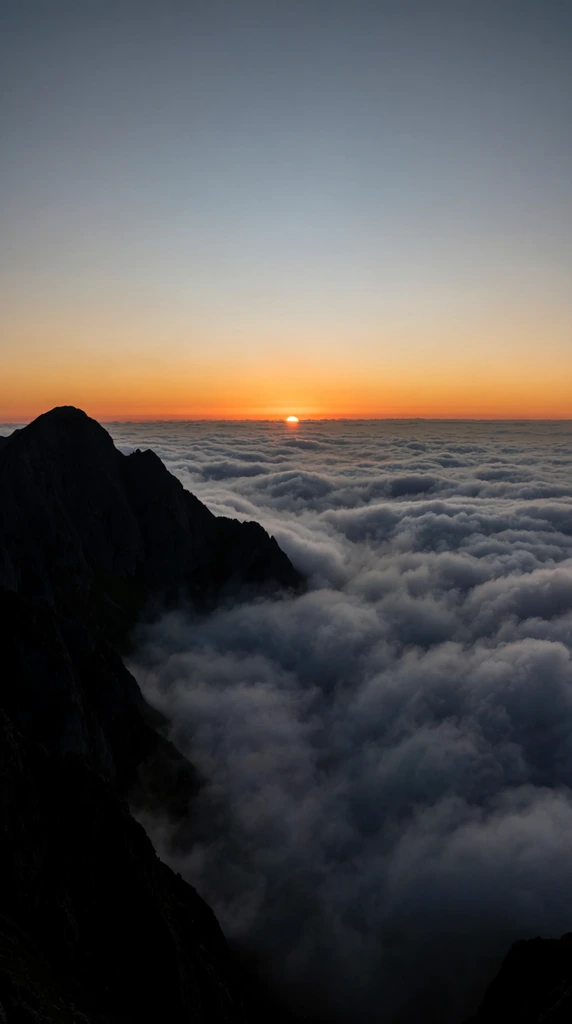 Sunrise over a vast sea of rolling clouds, dark jagged mountain silhouette dominating the left foreground, glowing orange sun breaching the distant cloud horizon, dense textured gray and white cloud inversion, smooth gradient sky shifting from muted blue to vibrant orange, sweeping high-altitude vista, dramatic landscape photography, high contrast