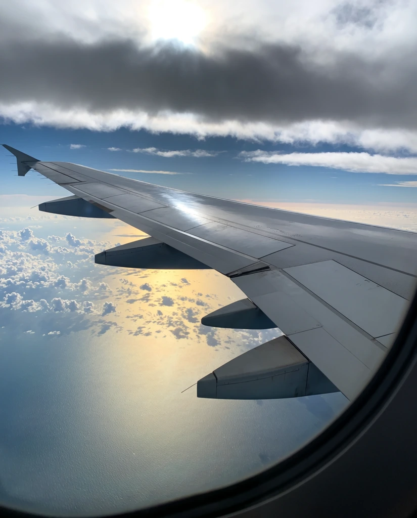 metallic grey airplane wing extending diagonally, aerodynamic flap fairings beneath the wing surface, curved black window frame at bottom right, high altitude aerial perspective, dark horizontal cloud band across the upper sky, patches of blue sky above, scattered fluffy cumulus clouds below the wing, sparkling ocean water far below, brilliant golden sunlight reflecting off the water surface, natural dramatic lighting, high contrast between mechanical aircraft structures and soft atmospheric layers
