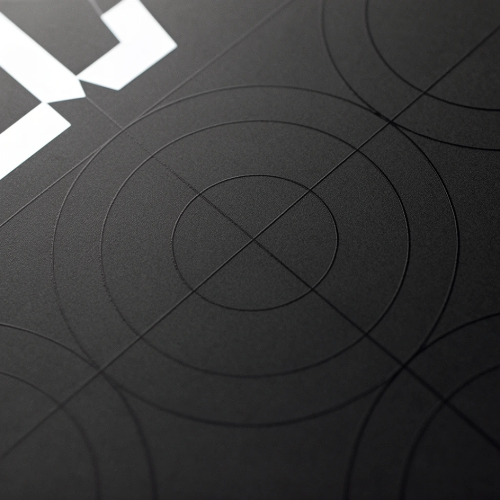 Precise geometric line art is etched into a dark charcoal, finely textured matte surface in this macro photograph. The minimalist composition features large concentric circles intersected by faint straight axes, resembling a clean technical diagram. A crisp, bright white abstract geometric shape sits in the upper left quadrant, providing stark contrast against the subtle dark-on-dark linework. Soft, directional illumination from the top left casts a smooth gradient across the grainy material, highlighting the slightly debossed nature of the circles. An oblique camera angle and shallow depth of field gently blur the bottom right edge of the frame, enhancing the sleek, premium industrial aesthetic of the abstract layout.