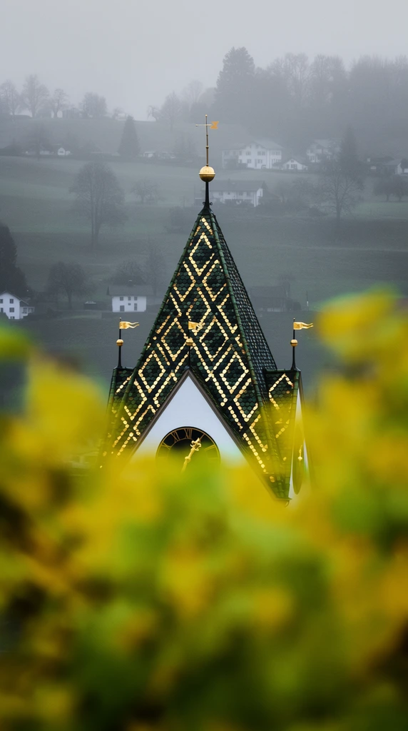 A sharply focused architectural tower with a steep, pyramidal roof emerges through heavily blurred, vibrant yellow-green foreground foliage. The roof features a diamond pattern of dark green and reflective golden tiles, topped by a golden sphere, a weather vane, and a cross finial. Small golden flag ornaments line the roof edge above a white facade partially revealing a dark clock face with golden hands. The out-of-focus foreground leaves create a dominant framing effect, obscuring the building's lower half. Behind the tower, a moody, misty rural landscape features rolling grey-green hills. Scattered white houses and softly silhouetted trees fade into the dense atmospheric fog, highlighting the shallow depth of field and compressed telephoto perspective.