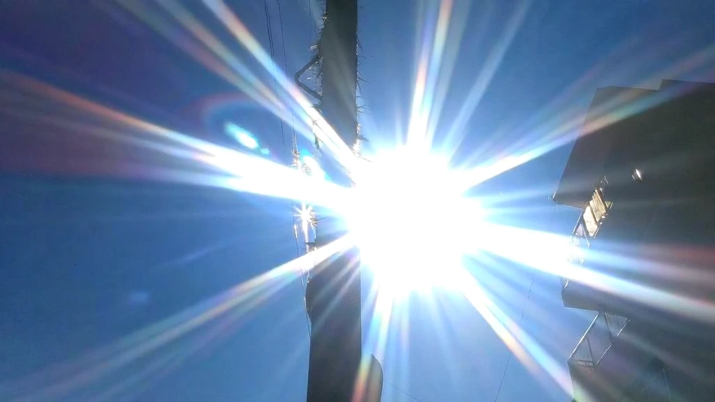 street intersection with a crosswalk, central utility pole, buildings with balconies on the right, low-angle shot looking upward, blindingly intense contra-jour sunlight, massive multi-pointed starburst lens flare with sharp radial rays, prismatic chromatic aberration on light edges, deep cerulean sky gradient, high-contrast silhouettes, overexposed highlight bleed, digital sensor bloom, hazy atmospheric glow, lo-fi smartphone photography aesthetic