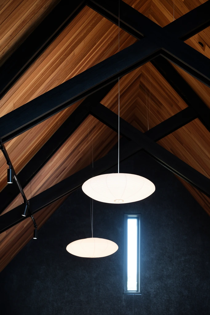 A low-angle architectural view of a pitched wooden ceiling with heavy, intersecting black structural beams and two glowing white ribbed pendant lights. Warm wood planks line the roof, contrasting sharply with the thick dark diagonal trusses. Suspended by thin wires, the squat, paper-like lanterns provide soft, luminous curves against the rigid geometry. Black track lighting is mounted on a left beam. The background features a textured dark charcoal wall with a single narrow vertical window radiating bright daylight, highlighting the dramatic interplay of heavy timber and delicate illumination.
