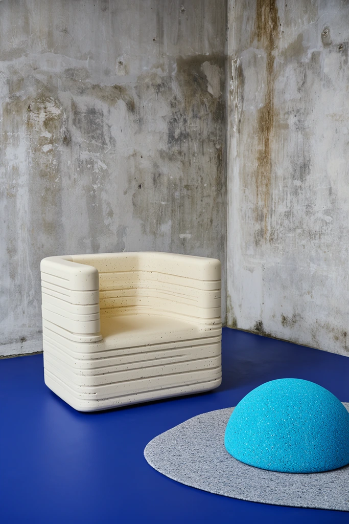 chunky cream-colored architectural lounge chair, stacked horizontal texture with linear pitting, raw concrete corner walls with mottled grey plaster and prominent stains, smooth solid royal blue floor, vibrant textured cyan dome resting on an irregular speckled grey mat, soft directional daylight, subtle ambient shadows, raw industrial textures contrasted with vivid solid colors, contemporary sculptural design