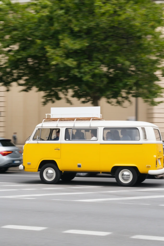 A vibrant yellow and white vintage microbus speeds down an urban street, captured with a dynamic panning technique. The two-tone van features a white roof and window pillars, a bright yellow lower body, chrome hubcaps, and a wooden roof rack holding a white rectangular board. Silhouettes of passengers are visible through the side windows. The van travels across a grey asphalt road with white painted markings. Horizontal motion blur softens the background, which includes a large leafy green tree, a beige building facade, and a modern silver car trailing on the left. The daytime photograph uses a slow shutter speed to blur the spinning wheels and static environment, keeping the main subject sharply in focus to emphasize its retro color palette.