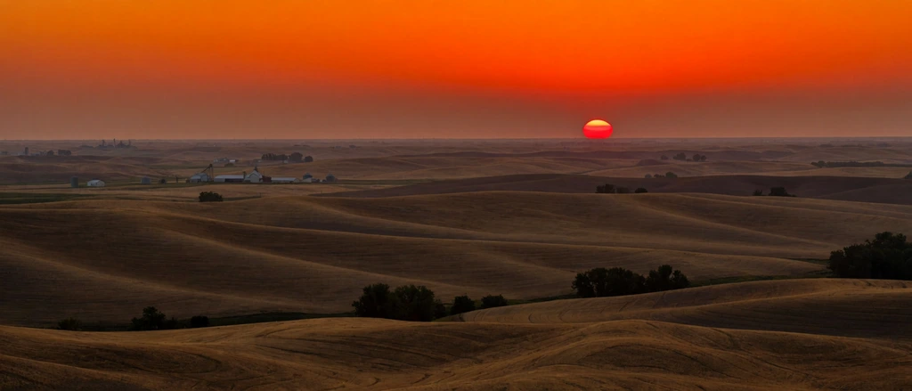 fiery orange sunset over vast undulating agricultural hills, glowing red half-sun on the flat horizon, deep shadows emphasizing rolling golden-brown terrain, scattered dark tree clusters, distant white farm buildings and grain silos nestled in shallow valleys, sweeping wide landscape photography, dramatic contrast, atmospheric haze