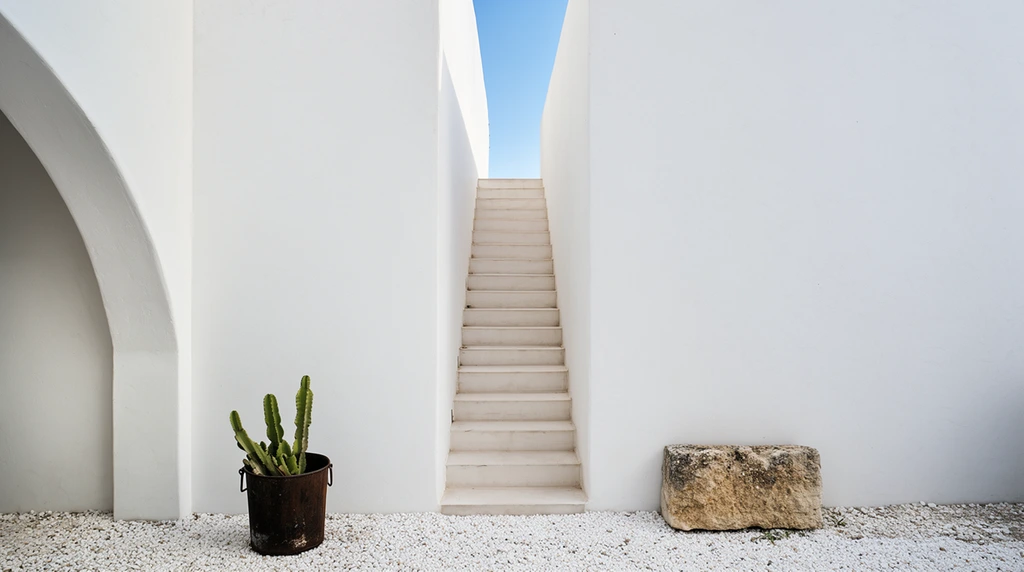 minimalist white architectural exterior with a central staircase ascending to a clear blue sky, straight narrow flight of pale steps flanked by towering stark white walls, partial structural arch on the left wall, green cactus in a weathered dark metal bucket on white gravel, rustic stone block on the right, bright natural daylight, architectural photography, minimalist composition, stark geometry, clean vertical lines, soft shadows