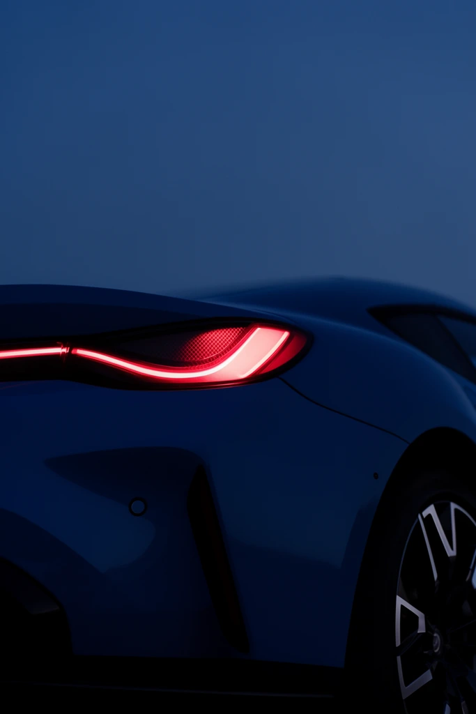 A close-up rear quarter view of a sleek dark blue sports car against a smooth twilight sky. Draped in deep shadows, the aerodynamic curves blend into the minimalist background. A glowing red taillight forms a sharp, illuminated halo with a textured inner mesh, creating a vibrant focal point. Below the light, a dark angular vent and a small circular sensor detail the bumper. A partially cropped black alloy wheel with geometric silver spokes occupies the lower right corner. The low-angle composition uses extensive negative space, capturing a moody, high-end automotive photography aesthetic defined by a monochromatic deep blue palette pierced by stark neon red.