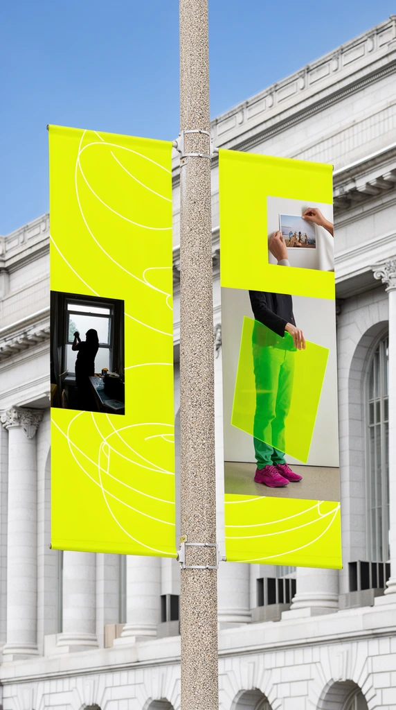 Two vibrant neon yellow vertical banners hang from a speckled concrete pole, set against a stately light grey stone building with classical columns and arched windows under a clear blue sky. The contemporary banners feature inset photographic squares and abstract white curving line art. The left banner includes a photo of a silhouetted figure working by a window. The right banner showcases a person wearing green pants and magenta sneakers holding a large translucent neon green rectangular panel, alongside an inset of hands pinning up a picture. Captured from a low angle in bright daylight, the composition highlights a striking juxtaposition between ornate historic architectural details and bold modern design elements.