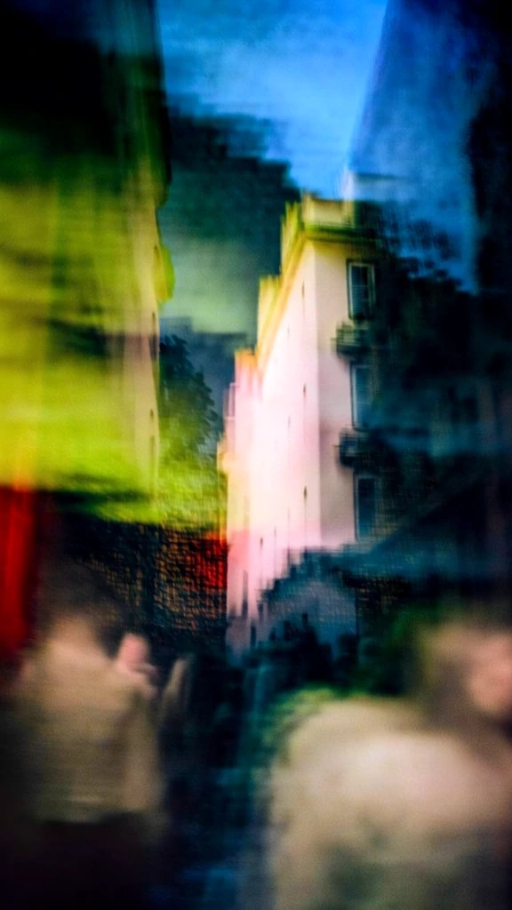 cinematic street scene with tall buildings flanking a crowded thoroughfare, heavy motion blur, double exposure ghosting effect with layered architectural lines, saturated palette of acid lime green and deep crimson accents against dark charcoal and cerulean shadows, soft painterly textures, ethereal glowing highlights, dreamlike long exposure aesthetic, high contrast shadows, hazy atmosphere, surreal superposition of urban facades and crowds, vibrant, moody, impressionistic style