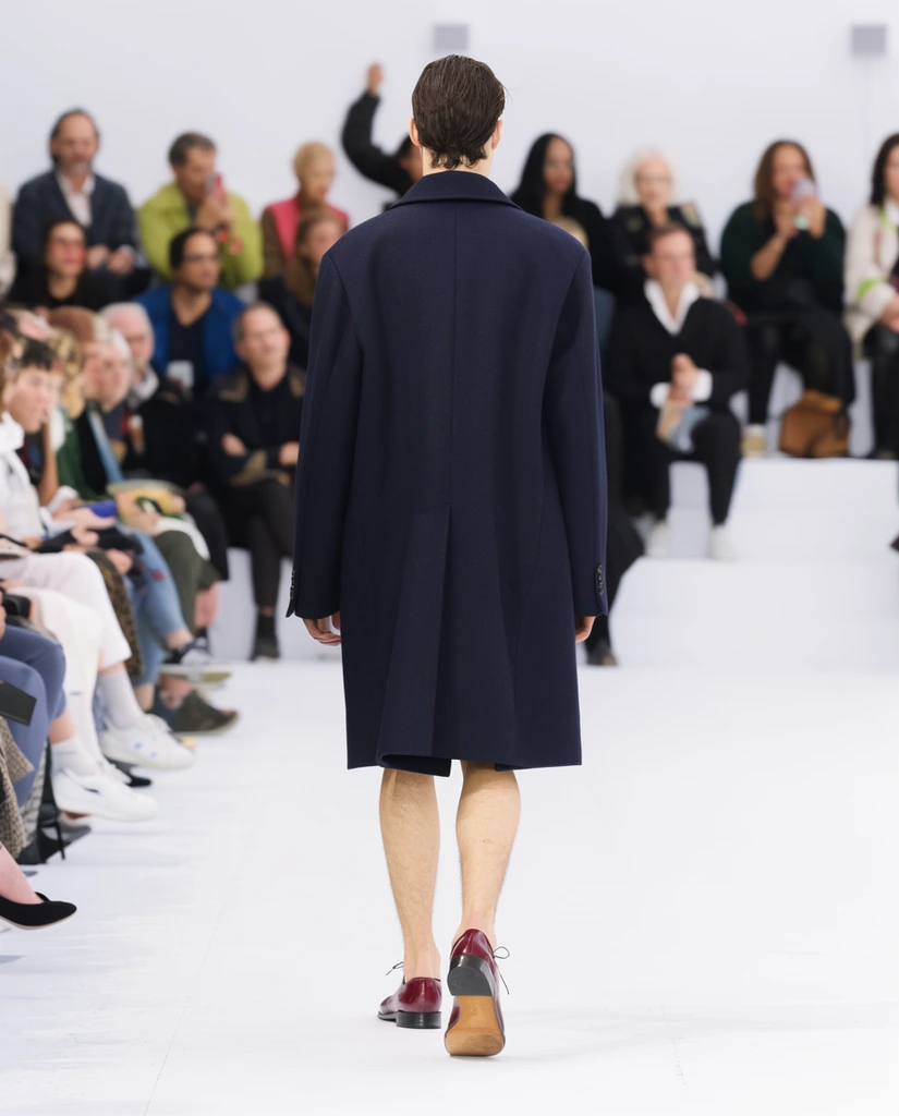 back view of model in oversized navy tailored coat, bare legs, maroon dress shoes, bright white runway, blurred seated audience, shallow depth of field, bright diffuse lighting