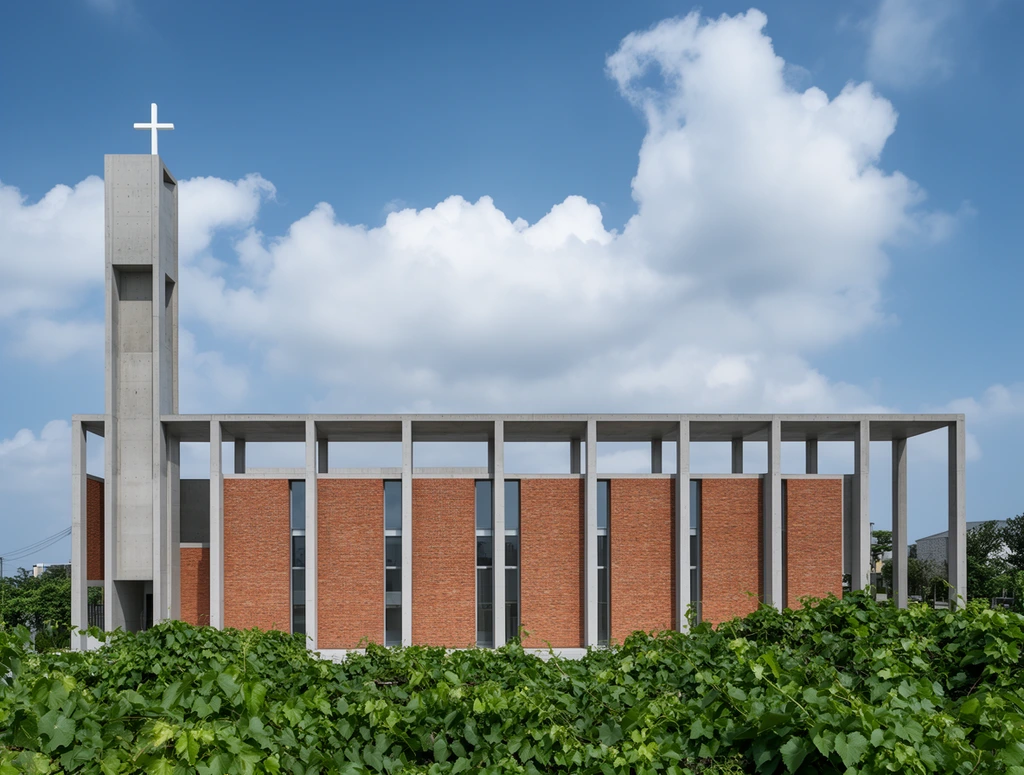 modern minimalist church exterior, exposed concrete and red brick facade, tall slender concrete tower topped with a white cross, open flat-roofed colonnade, recessed glass windows, dense foreground of lush green vines, bright daylight, blue sky with fluffy cumulus clouds, crisp geometric lines, architectural photography