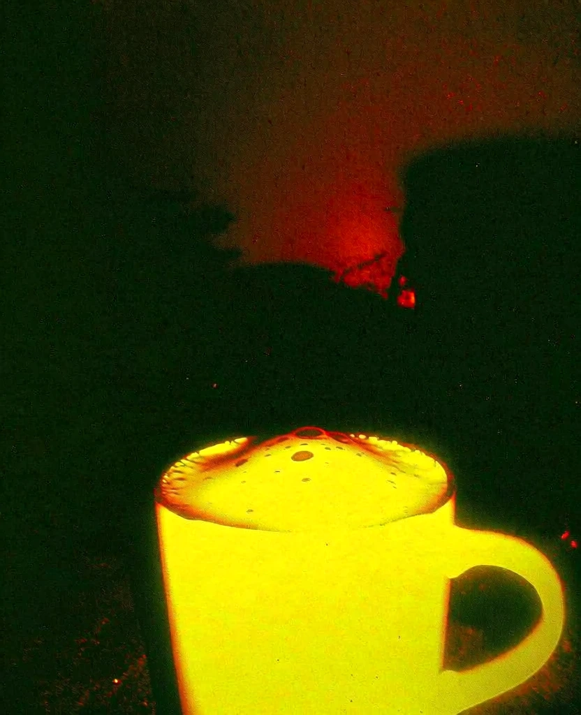low-angle close-up of a mug filled with a caffeinated drink, thick frothy foam with bubbles of various sizes, extreme high-contrast tri-tone aesthetic, mug glowing in flat radioactive neon yellow with blown-out highlights, deep pitch-black crushed shadows, background features a hazy blood-red and burnt-orange glow behind dark silhouettes, heavy analog film grain, gritty texture with dust specks and noise, lo-fi experimental photography style, thermal imaging palette, eerie atmosphere.