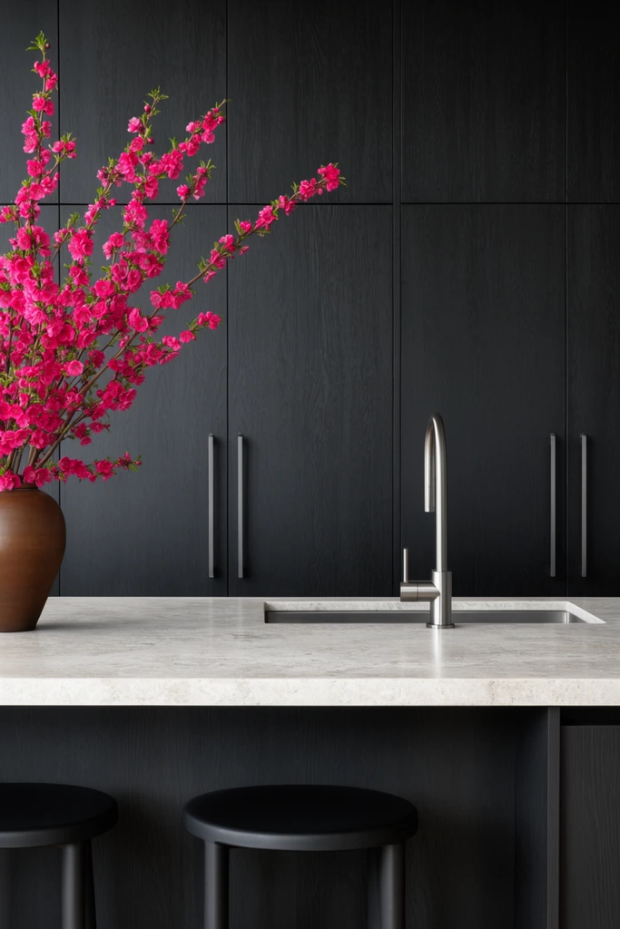 A minimalist kitchen featuring black wood grain cabinetry and a light stone island accented by vibrant pink blossoms. The floor-to-ceiling dark cabinets feature slim vertical steel pulls. A light grey textured countertop holds a sleek steel gooseneck faucet over an undermount sink, with matte black stools tucked beneath the overhang. A brown ceramic vase displays branches dense with bright magenta flowers, creating a stark organic contrast against the austere architectural geometry under soft diffused lighting.