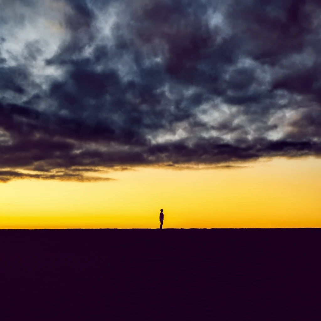 A lone, silhouetted figure stands on a dark, flat horizon line against a dramatic sky. The horizon sharply divides the image horizontally, with the lower third consisting of a featureless, deep purple-black foreground. Above the horizon, a brilliant, glowing yellow-orange band of sunset sky stretches across the width. Above this luminous band, an expansive, heavy canopy of dark, textured clouds dominates the upper two-thirds of the image, rendered in deep blues, grays, and purples. The clouds are dense, with swirling patterns and varying shades of darkness, creating a stark contrast against the bright lower sky. The tiny silhouette of the person is positioned near the center of the horizon, facing away or sideways, emphasizing the vast scale of the landscape and sky. The composition is minimalist, relying on strong horizontal lines and stark color contrasts to create a sense of immensity and isolation.