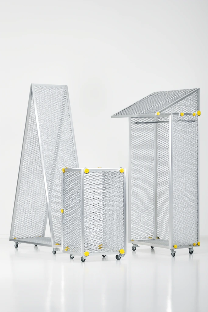 Three minimalist industrial structures built from silver expanded metal mesh and aluminum framing stand in a seamless white studio. The left piece forms a tall triangular A-frame on small caster wheels. Centered is a lower rectangular block accented with bright yellow circular fasteners at its joints. The right structure resembles a rolling open wardrobe with an overhanging mesh roof. The diamond-patterned grating creates a semi-transparent, moiré effect against the stark background. High-key diffused lighting casts soft reflections on the glossy floor, emphasizing the sleek metallic surfaces and sparse pops of yellow in the monochromatic environment.
