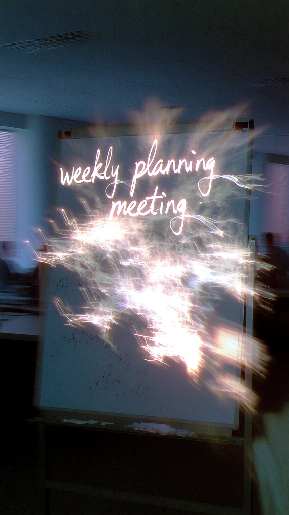 ultra-high-contrast flash photography, grainy analog texture, a whiteboard in a dark office with extreme motion blur, ghostly light trails emanating from the handwritten text weekly planning meeting across the top, harsh overexposed highlights on the whiteboard surface creating an ethereal glow, deep cyan and midnight blue shadows, blurred office silhouettes in the background, long exposure light painting, gritty cinematic realism, chaotic light streaks, raw lo-fi camera flash aesthetic