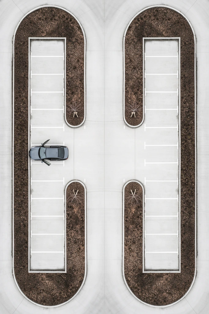 An aerial, top-down view of a highly symmetrical, geometric parking layout contrasting pale concrete with dark earth. A single dark sedan with an open driver-side door sits in one of the curved parking bays. Crisp white lines delineate spaces, creating a mirrored pattern across a central vertical lane. Four planters filled with textured brown soil—two large outer frames and two inner U-shapes holding small bare trees—form a graphic, abstract composition. The minimalist, desaturated color palette emphasizes precise architectural geometry and a stark aesthetic.