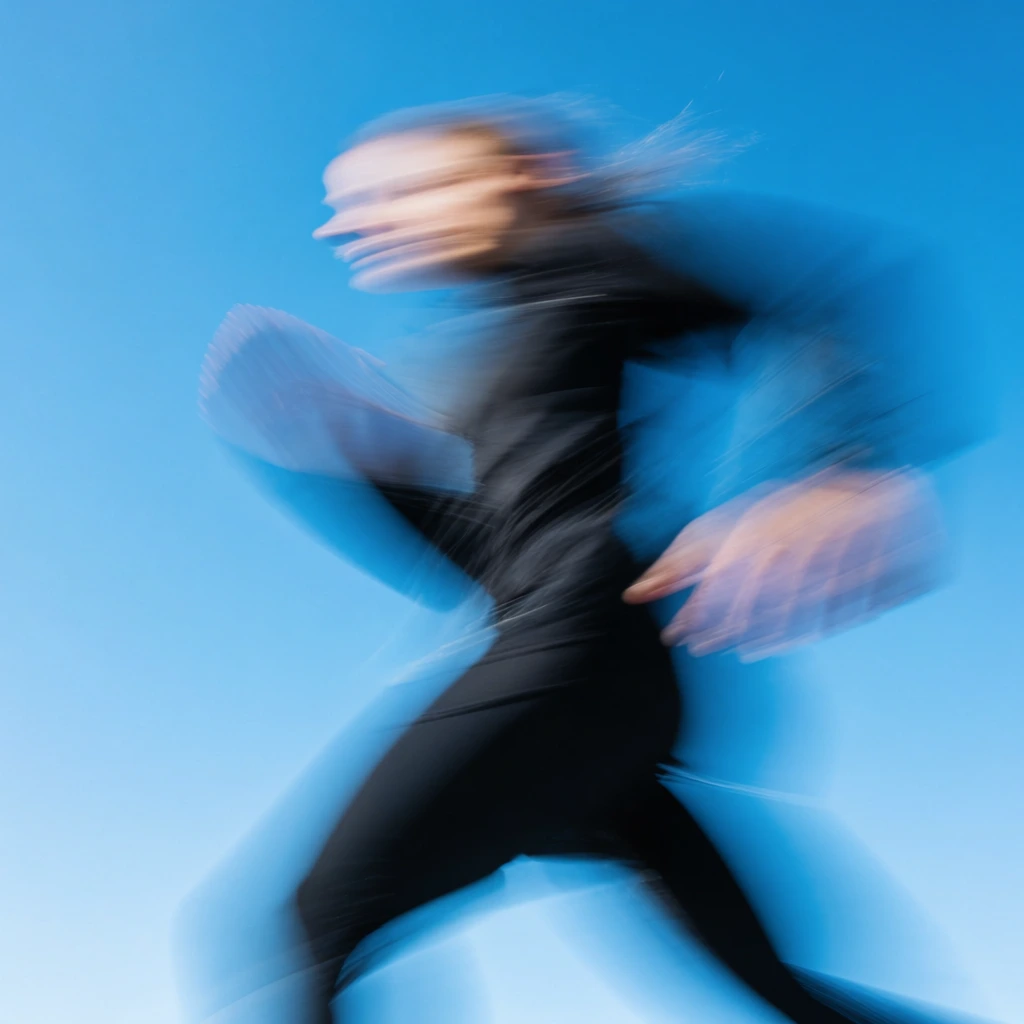 extreme motion blur photography of a person running, highly dynamic forward movement, heavily blurred figure wearing dark clothing, sharp contrast against a vibrant solid sky blue background, panning shot technique, high energy athletic speed effect, abstract streaks conveying velocity, indistinct light skin tone on the head, contemporary sports photography aesthetic, energetic atmosphere