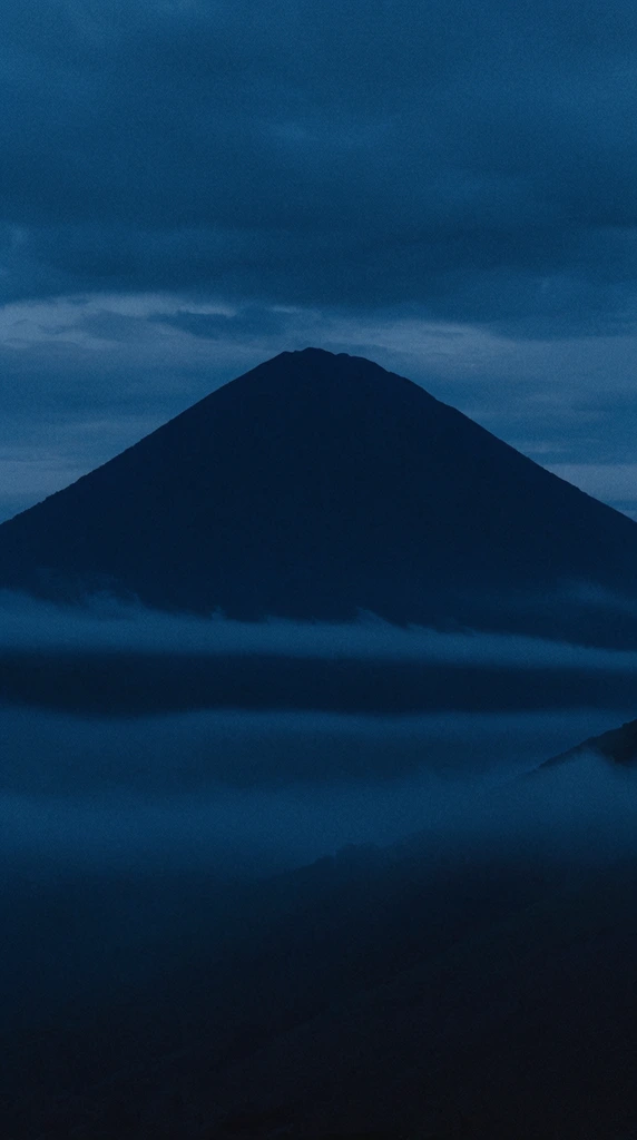 massive conical mountain silhouette towering at twilight, deep monochromatic navy blue color palette, thick horizontal bands of mist obscuring the lower slopes, dark atmospheric environment, overcast sky, cinematic landscape photography, filmic grain