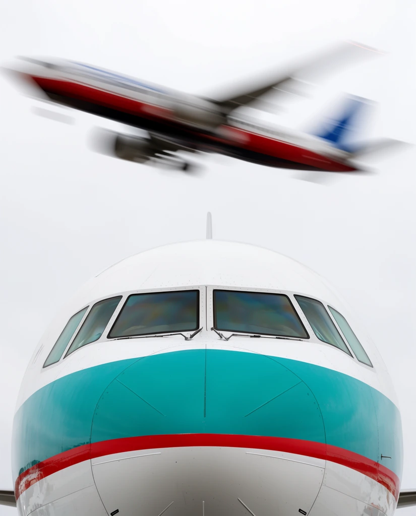 Close-up low-angle view of a commercial airplane nose and cockpit in sharp focus, featuring a white fuselage with a wide teal band and red stripe. Above it, against a uniform bright overcast sky, a second plane flies overhead, captured with strong motion blur showing streaks of white, red, and blue.