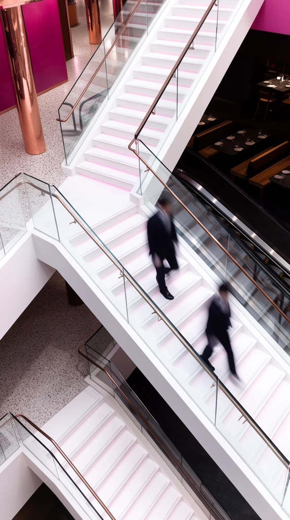 A high-angle architectural photograph captures a modern, multi-level staircase where two blurred figures in dark suits rapidly descend. The stark white stairs crisscross dramatically over dark voids, illuminated by a faint pink glow on the risers. Clear glass balustrades with bronze handrails frame the sharp, geometric steps. The surrounding interior features speckled terrazzo floors, reflective copper pillars, and vibrant magenta wall panels. To the right, a dimly lit dining area with dark tables and wooden benches sits beside an escalator. Strong diagonal lines dominate the composition, contrasting the highly structured, futuristic commercial environment with the long-exposure motion blur of the passing subjects.