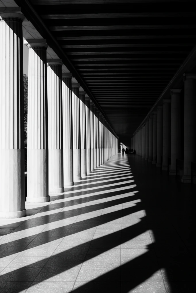 A high-contrast black and white photograph of a classical colonnade receding into a deep one-point perspective. Massive fluted columns on the left are struck by harsh sunlight, casting rhythmic, sharp diagonal shadows across a smooth tiled floor. A dark beamed ceiling and heavily shadowed columns on the right frame the expansive corridor, drawing the eye toward tiny silhouetted figures at the distant vanishing point.