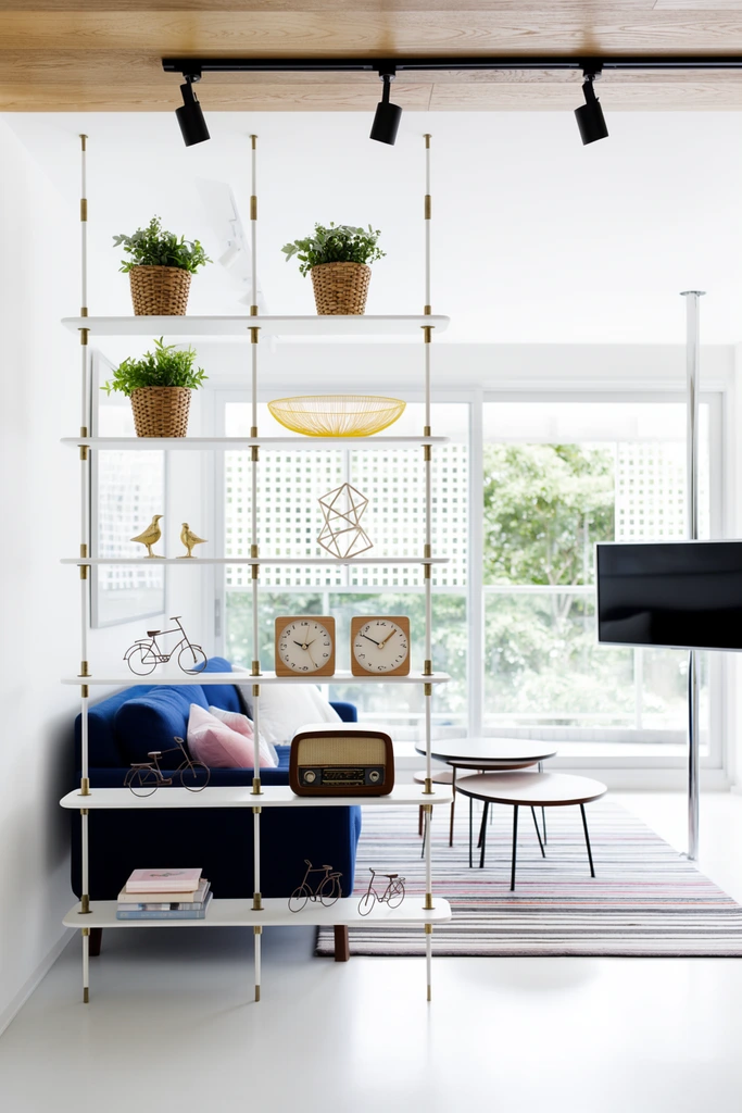 A floor-to-ceiling suspended white shelving unit with brass connectors serves as a room divider in a bright contemporary living room. The open shelves hold curated decor: potted plants in woven baskets, a yellow wire bowl, golden bird figurines, a geometric sculpture, square wooden clocks, a vintage wooden radio, miniature wire bicycles, and stacked books. Behind the divider sits a deep blue sofa with white and pink pillows alongside round nested coffee tables on a striped area rug. A flat-screen television mounts to a vertical chrome pole on the right. Large glass sliding doors in the background open to a balcony with white lattice screens and lush greenery, flooding the crisp white interior with natural daylight. Black track lights hang from a wood-paneled ceiling section directly above the shelves.
