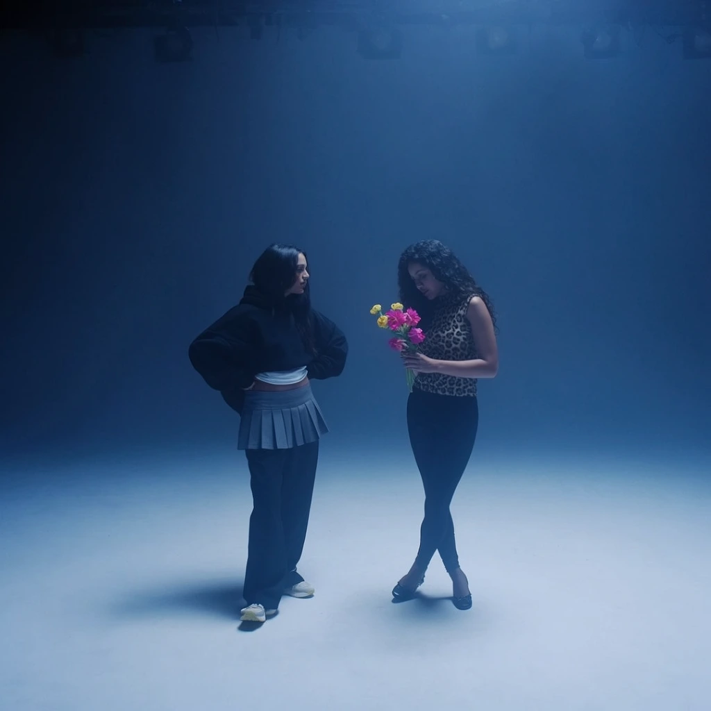 Two women stand in a hazy, expansive studio bathed in deep blue cinematic lighting. The woman on the left stands with hands on hips, looking at her companion, wearing an oversized black hoodie, a white top, a grey pleated mini skirt layered over loose black trousers, and pale sneakers. The woman on the right has long dark curly hair and looks down at a small bouquet of bright pink and yellow flowers. She wears a sleeveless leopard-print top, black leggings, and dark shoes with crossed ankles. They occupy a seamless cyclorama floor reflecting a soft spotlight, with faint silhouettes of overhead studio lights visible in the shadowy upper edge. The ethereal, minimal composition emphasizes cool tones and subtle atmospheric fog.